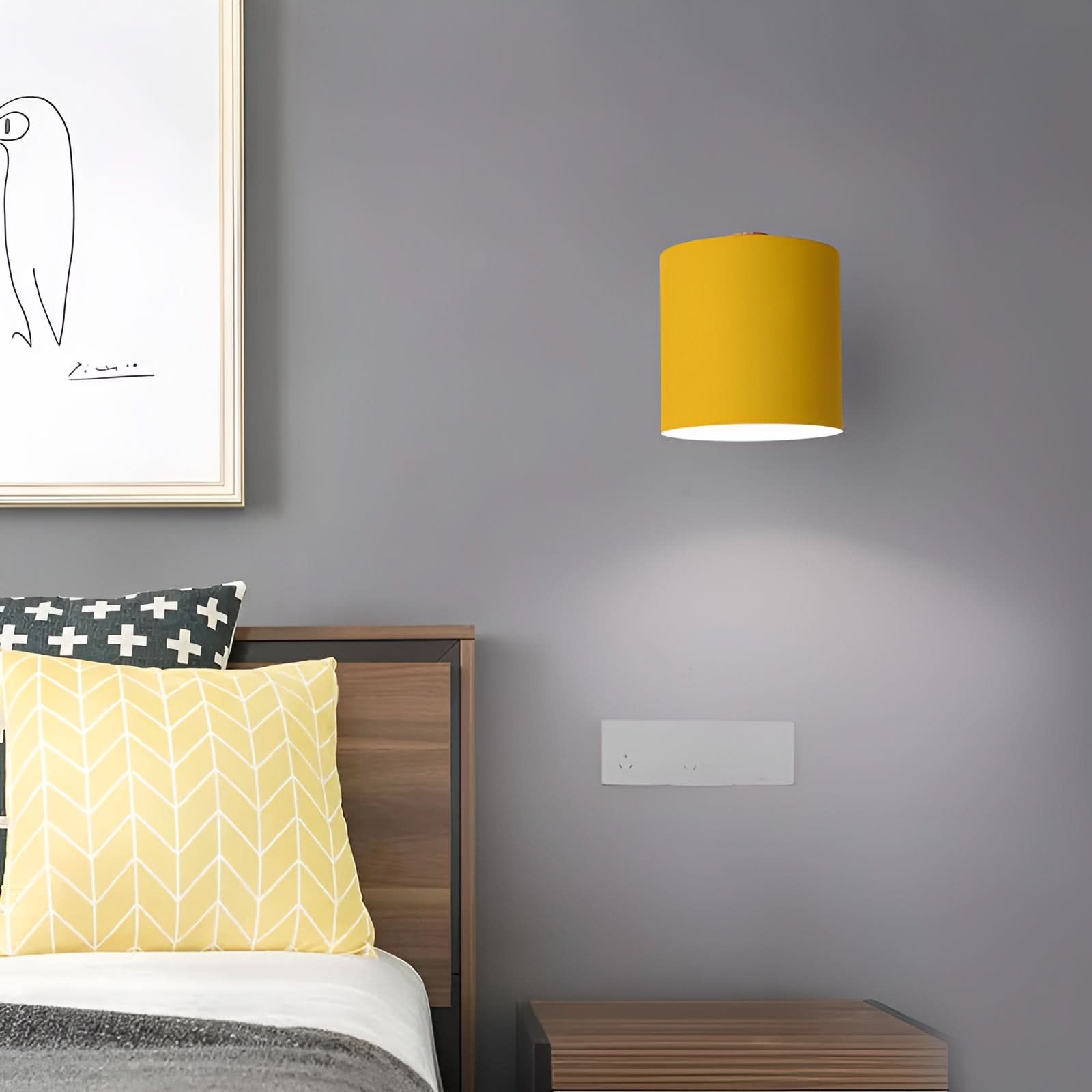 Yellow wall lamp on a gray wall above a bed with yellow and patterned pillows.
