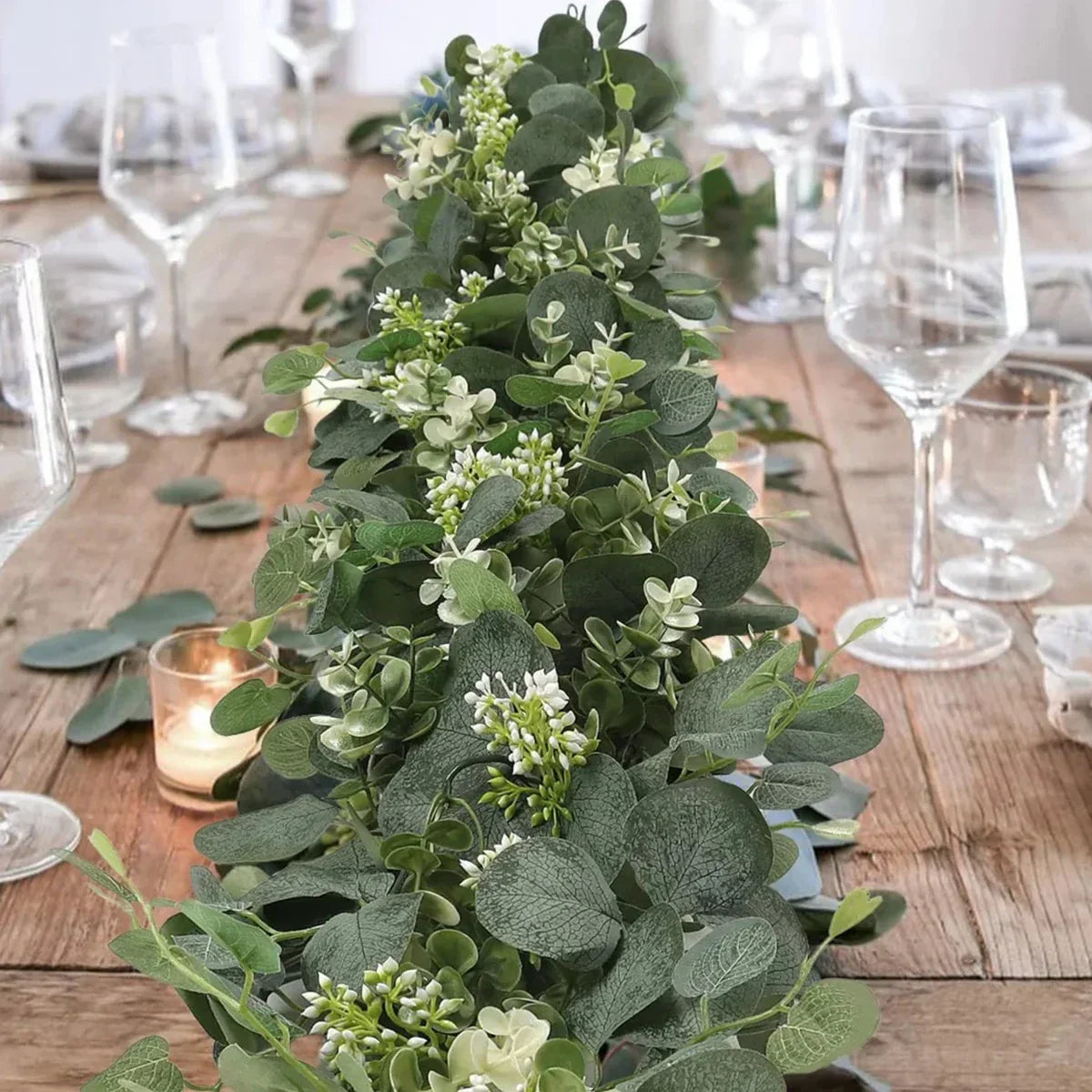 Decorative greenery arrangement on a wooden table with glasses and candles.