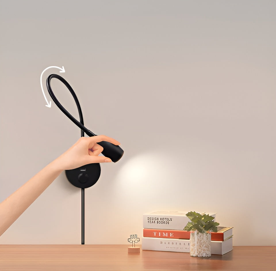 Person adjusting a black desk lamp on a wooden surface with books and a plant in the background.