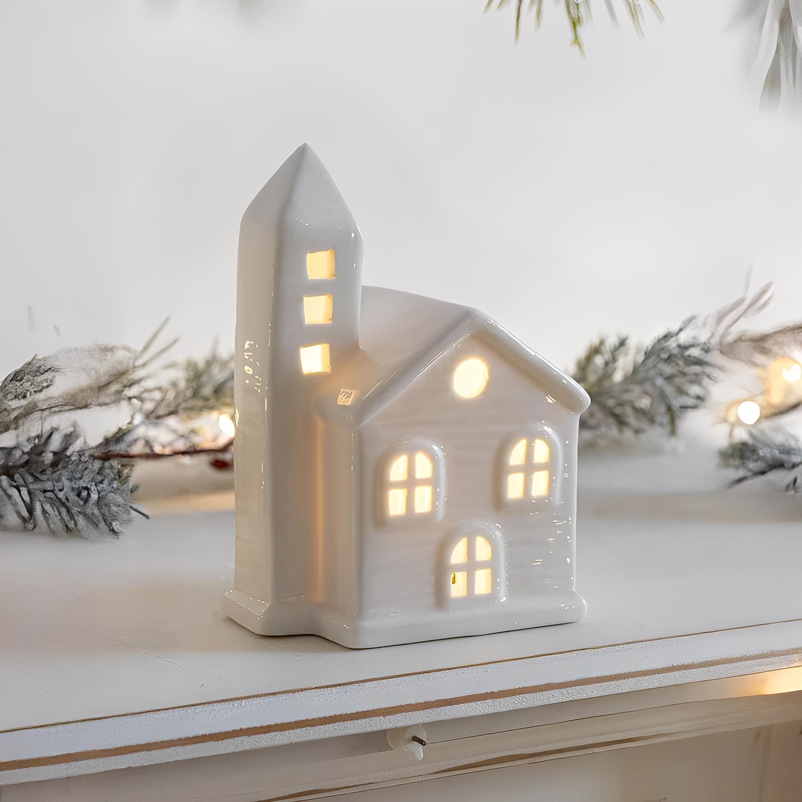 White ceramic house-shaped lantern with glowing interior on a white surface with decorative branches.