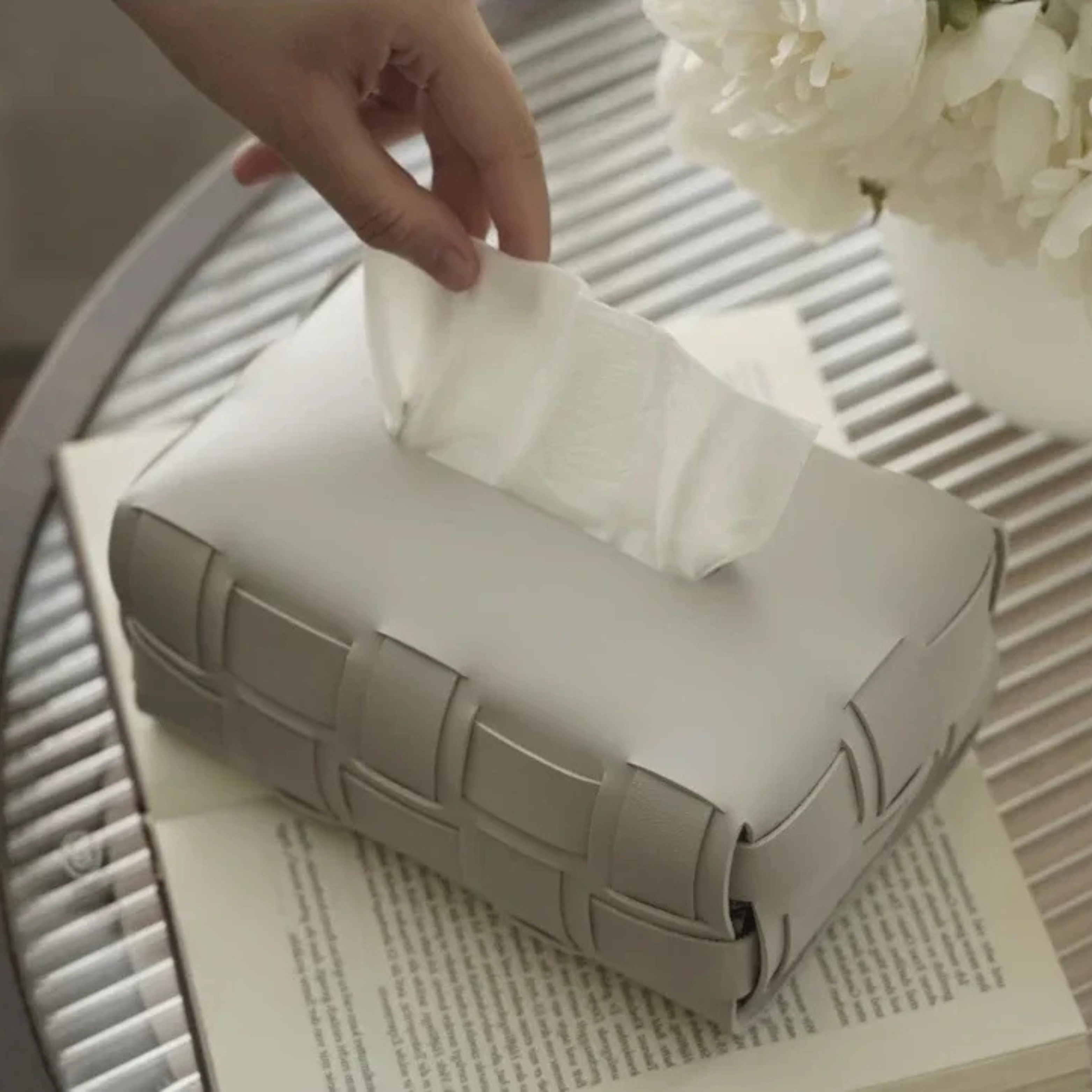Hand pulling a tissue from a woven tissue box on a table with flowers in the background