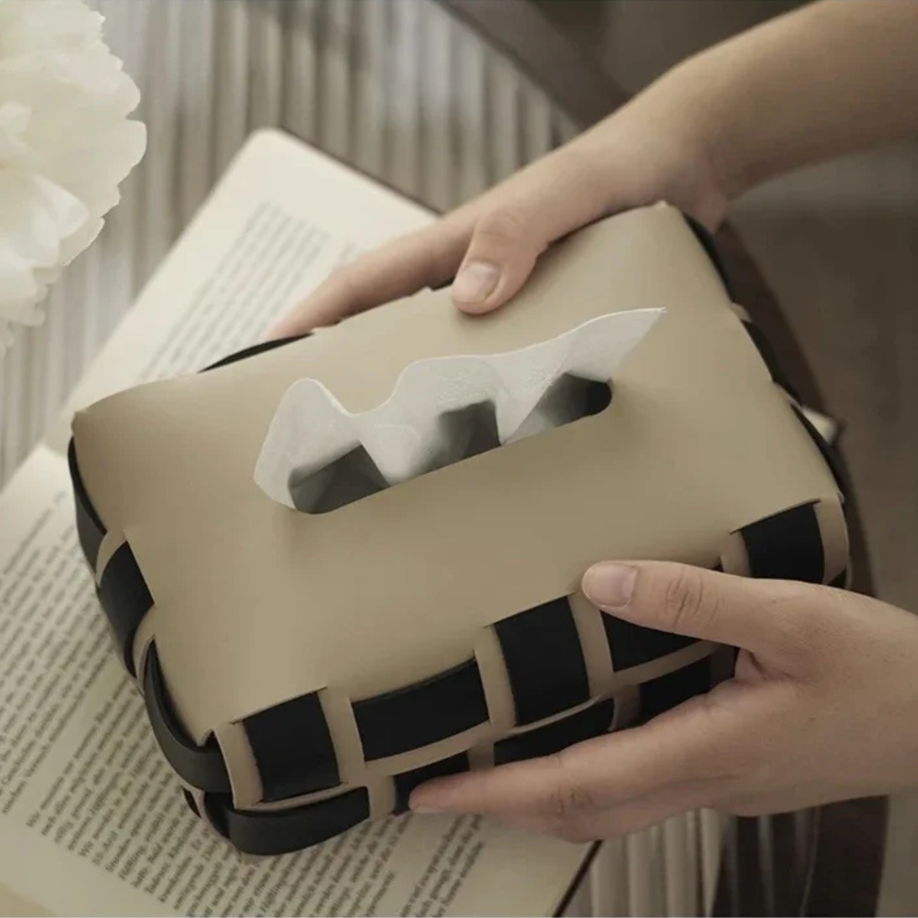 Beige and black woven tissue box held by hands on a surface with a blurred background