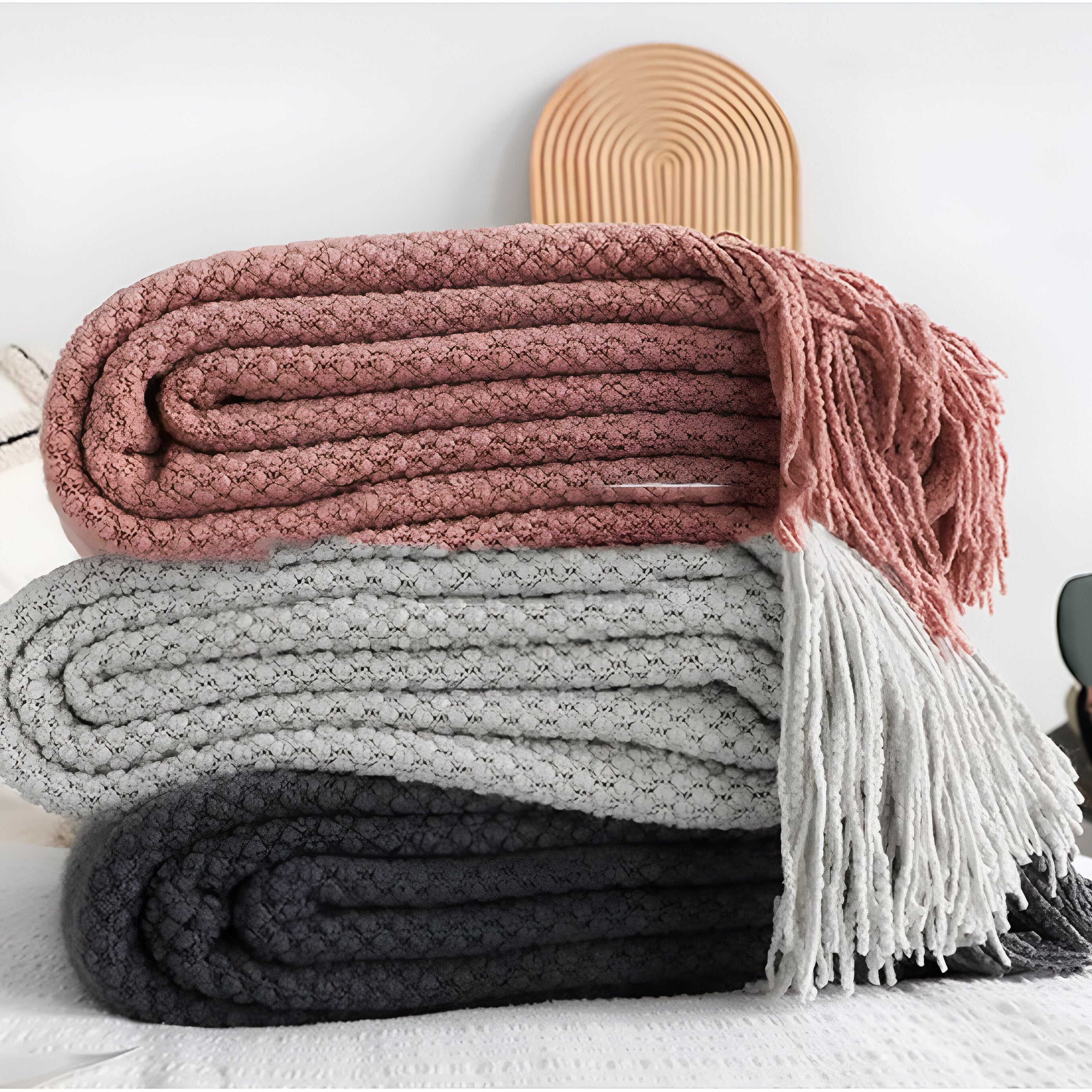 Stack of three folded blankets in pink, gray, and black with a tassel on a white surface.
