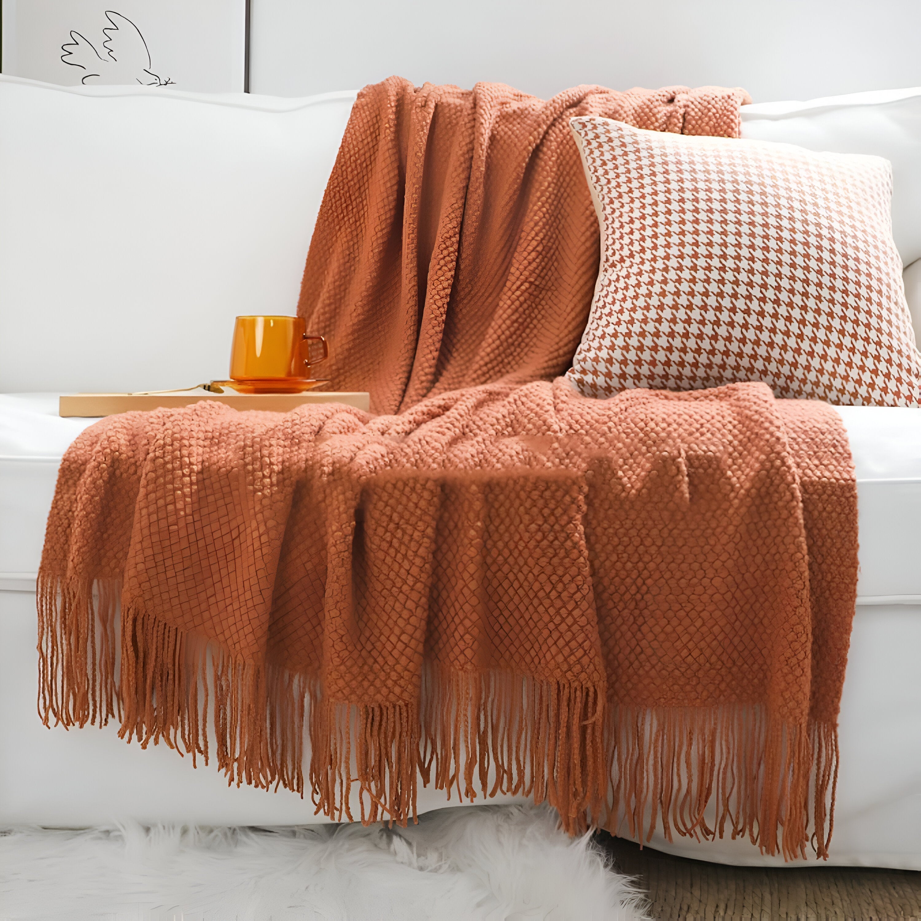 Orange throw blanket with fringe draped over a white sofa with a cup and tray on a wooden table.