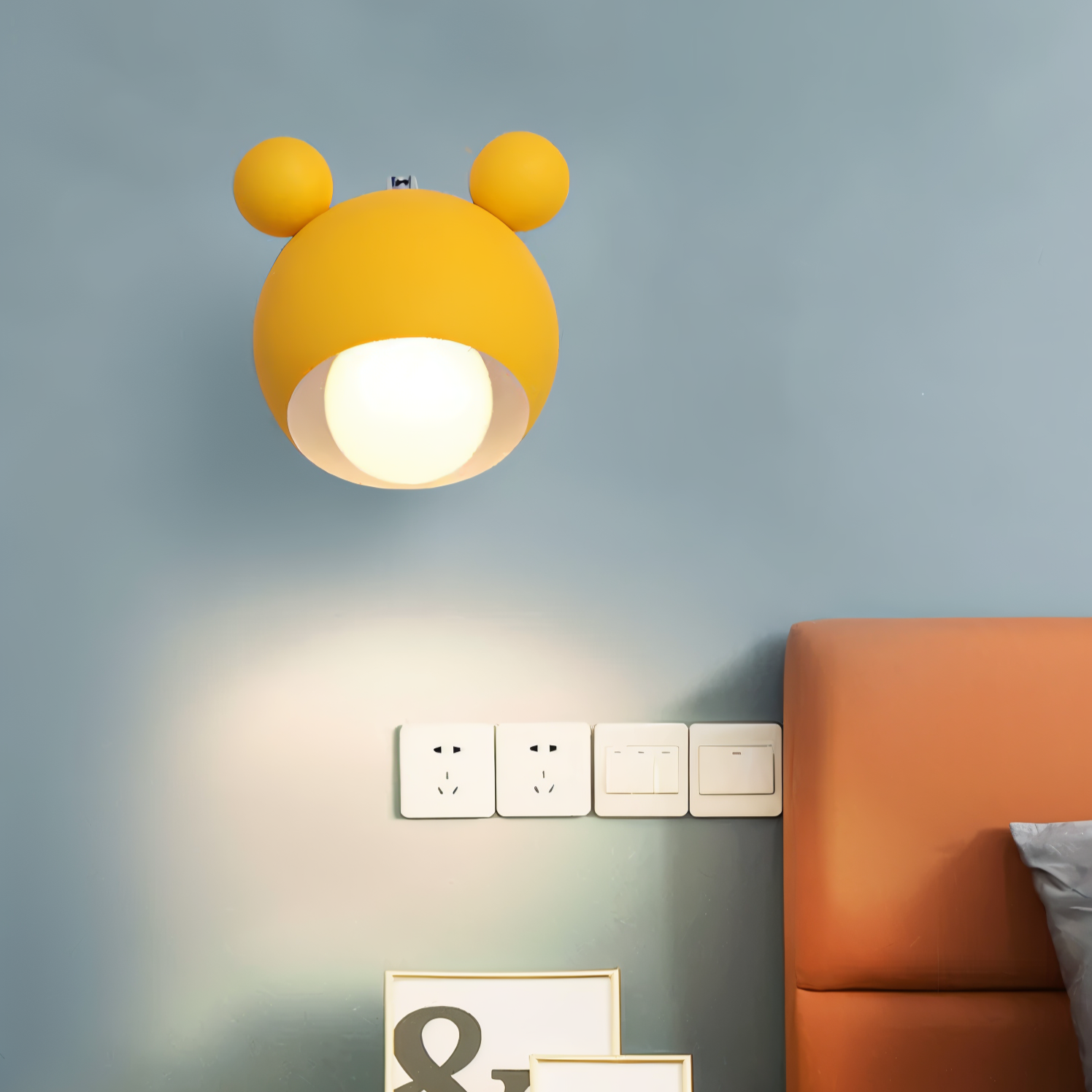 Yellow wall lamp with bear ears on a gray wall next to an orange sofa.