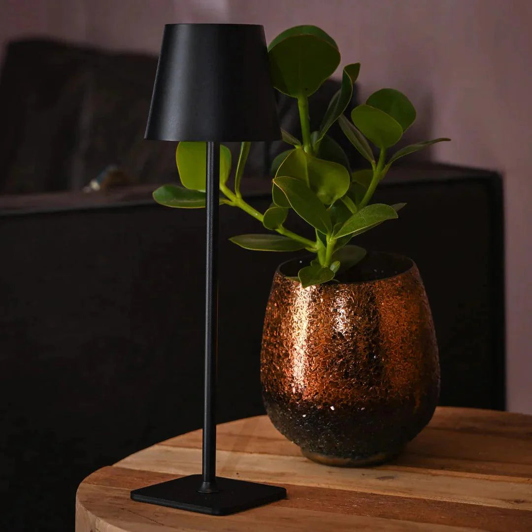 Black table lamp with a copper vase containing a plant on a wooden surface.