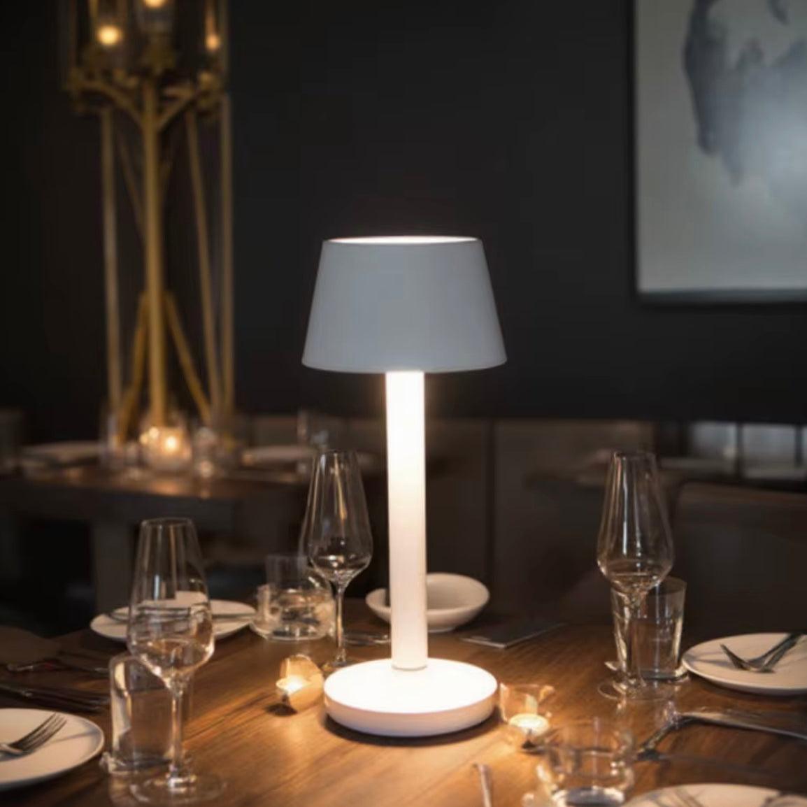 White table lamp on a wooden table with glasses and plates in a dimly lit room.