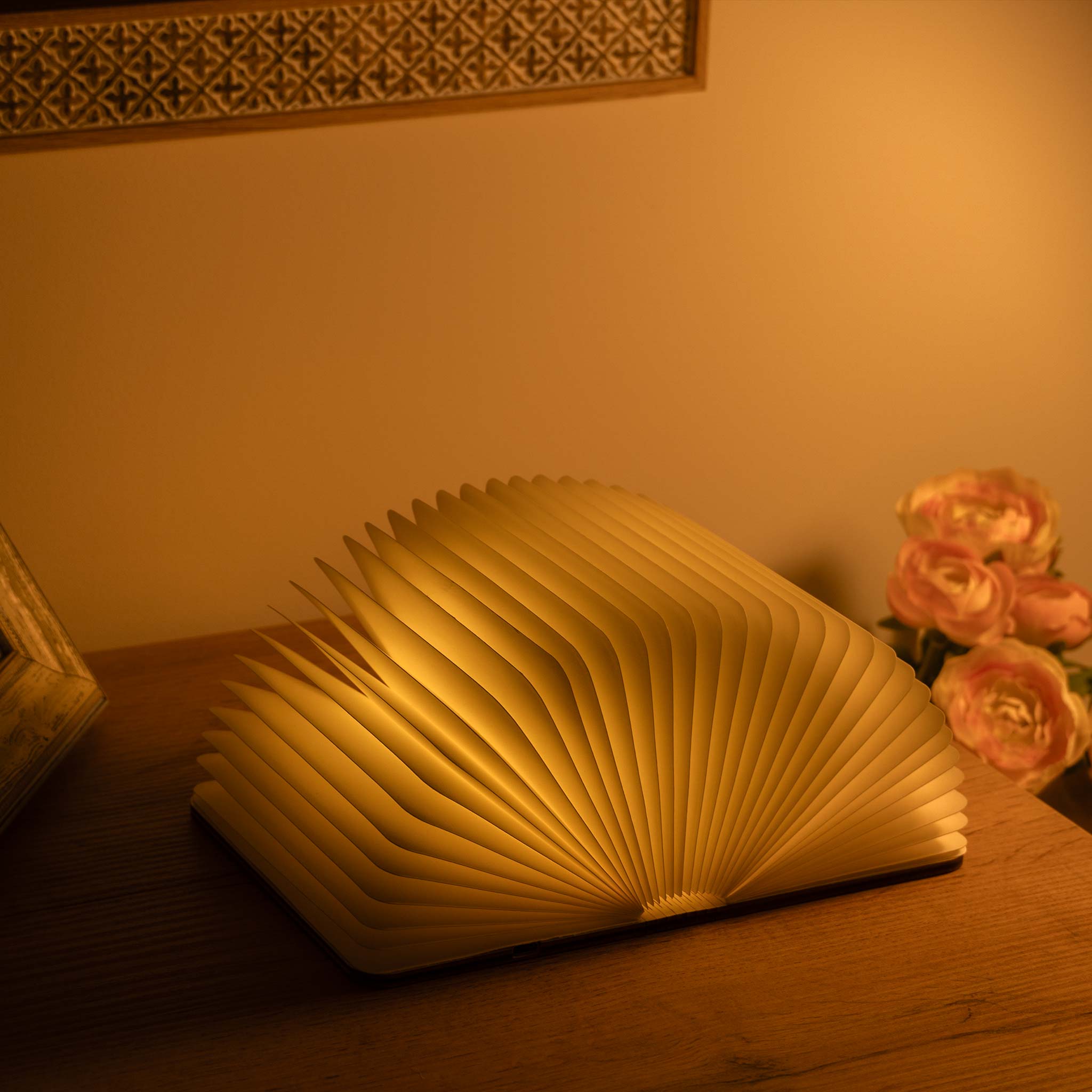 Book-shaped lamp emitting a warm glow on a wooden surface with flowers in the background.