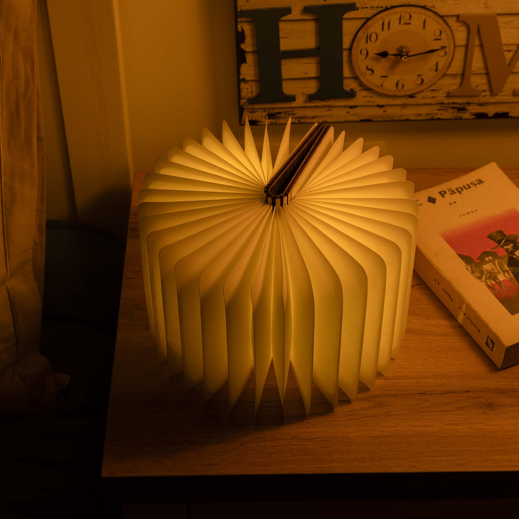 Book-shaped lamp on a wooden surface with a clock and book in the background