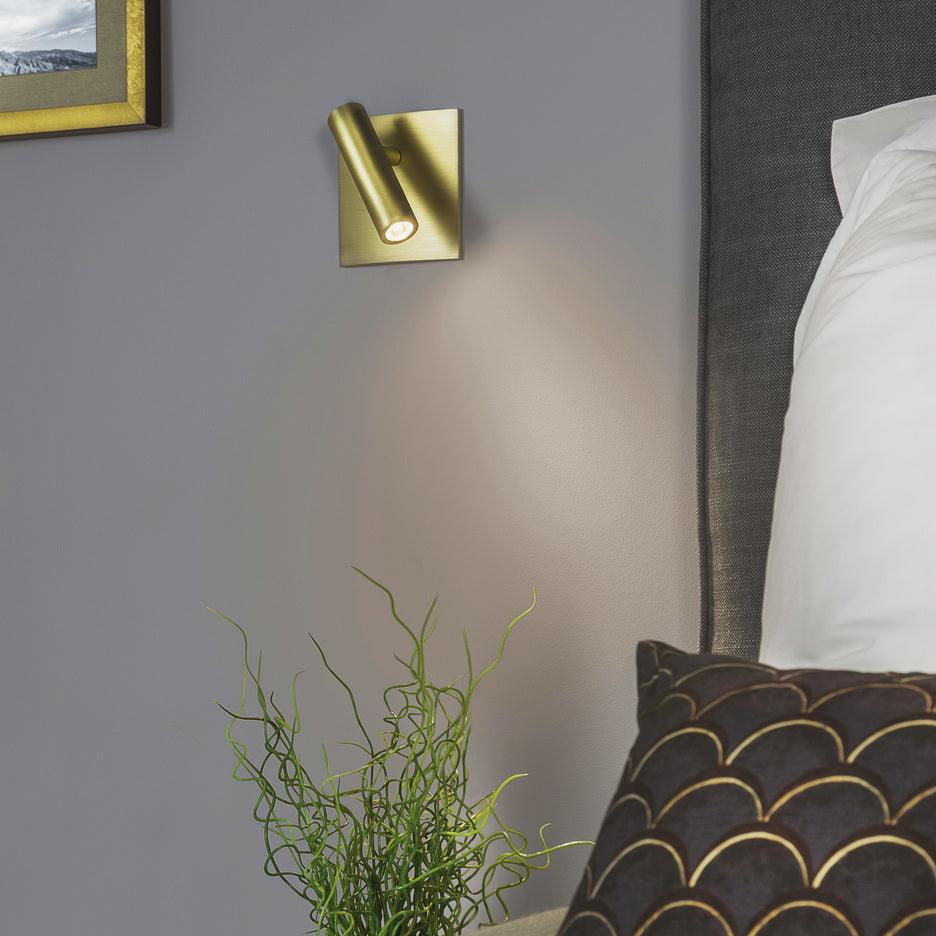 Gold wall sconce on a gray wall next to a bed with pillows and a plant.