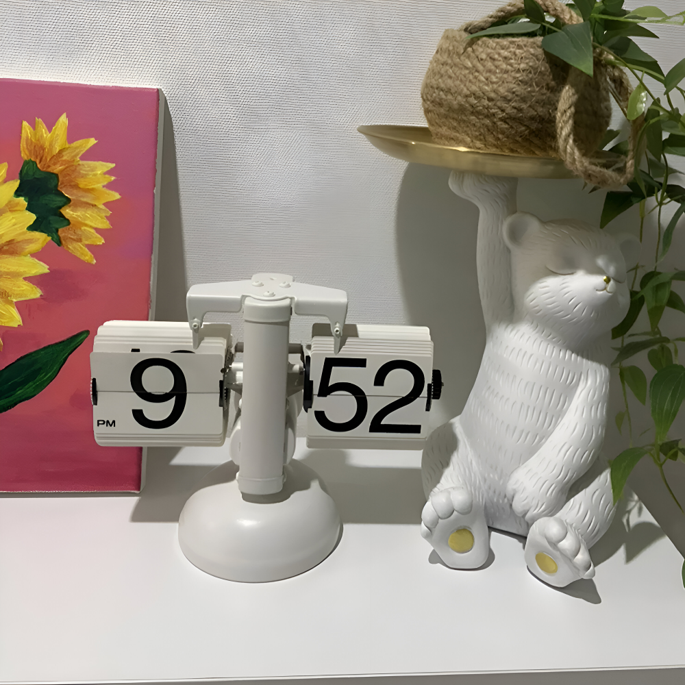 White clock with black numbers on a shelf next to a decorative bear and a painting of sunflowers.