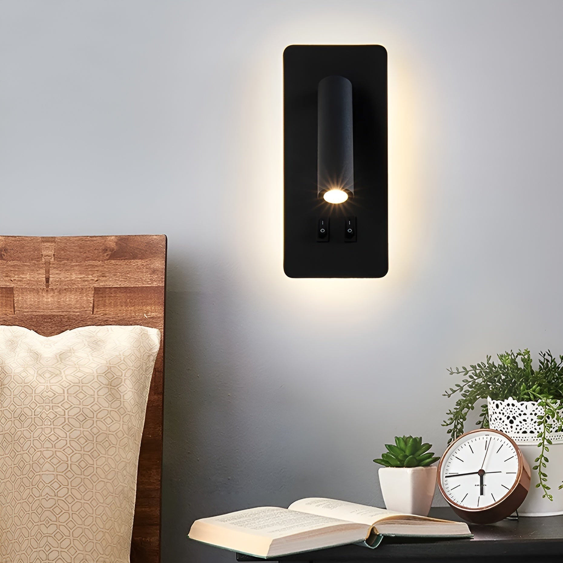 Modern wall sconce light fixture on a gray wall above a wooden headboard and nightstand with books and plants.