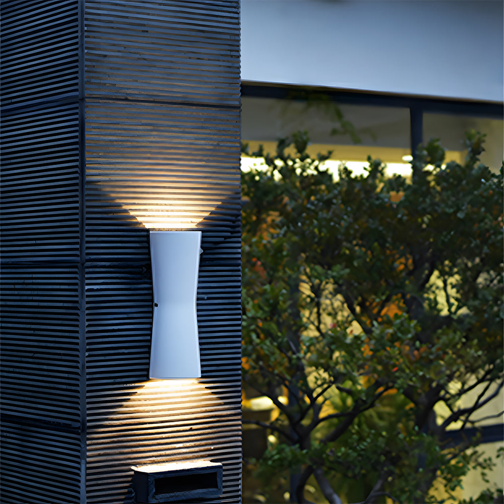 White outdoor wall light fixture on a textured stone wall with blurred and plant background