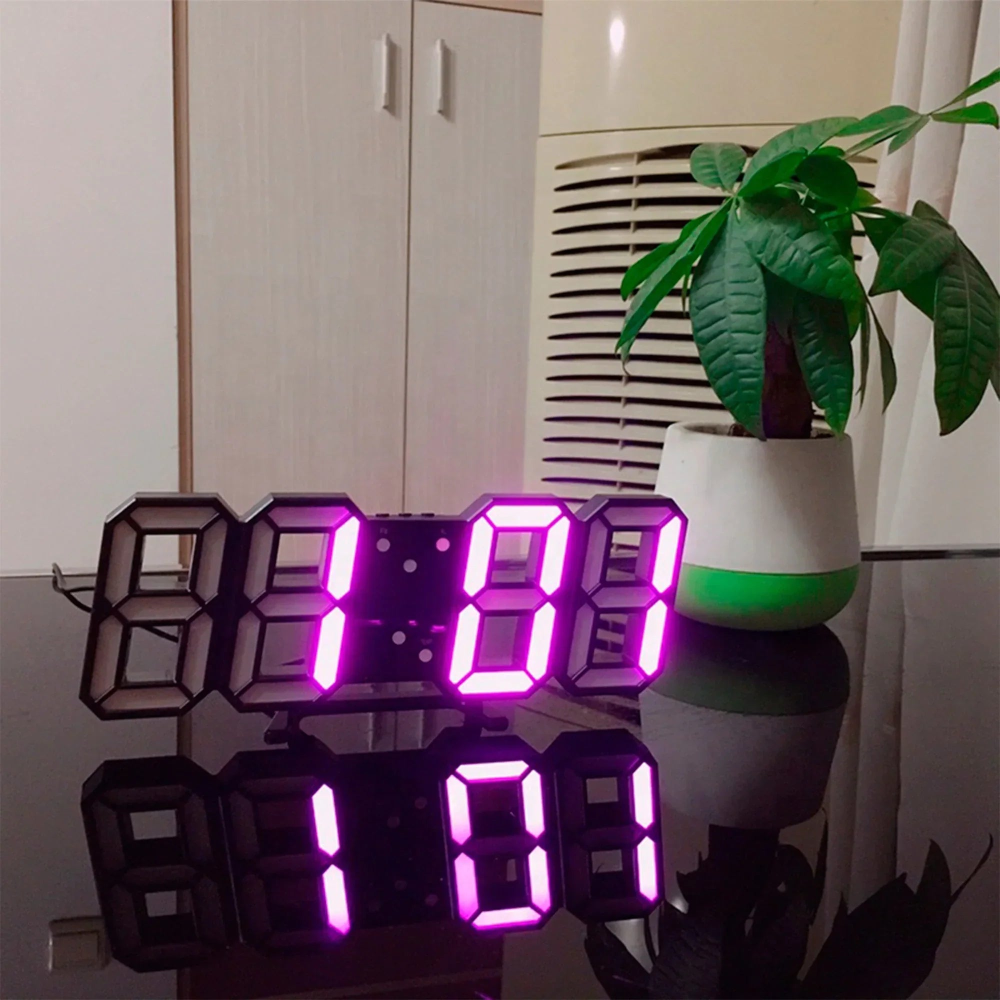Digital clock with purple LED display on a reflective surface, plant in the background