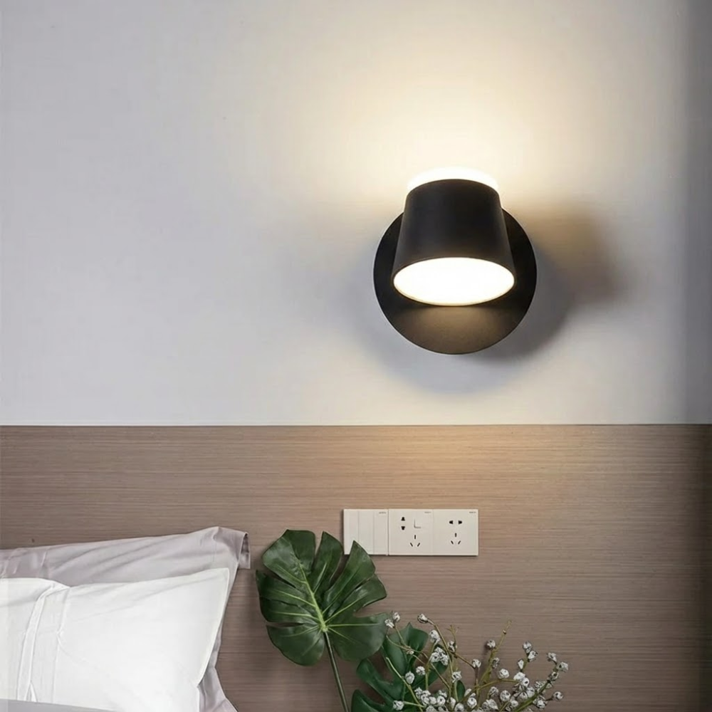 Modern wall-mounted light fixture on a neutral wall with a plant and bed in the foreground.