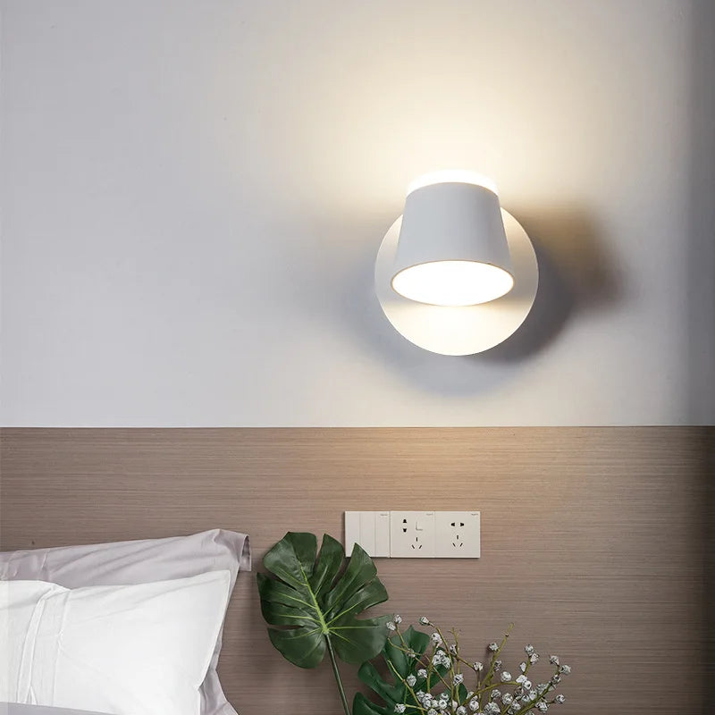 Modern wall-mounted light fixture on a neutral wall above a bed with a plant in the foreground.