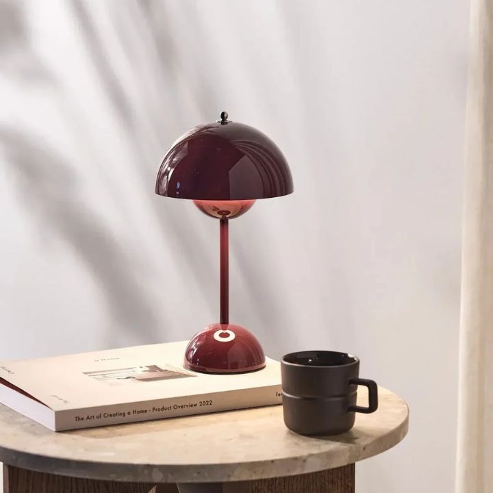Table lamp with a maroon shade on a table next to a book and black mug.