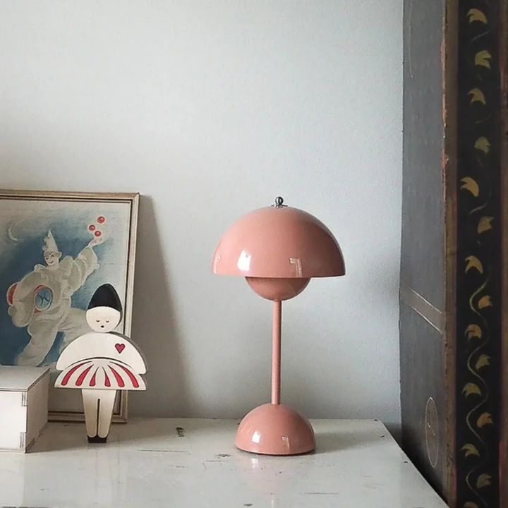 Pink table lamp on a white surface with a light gray wall and framed artwork in the background.