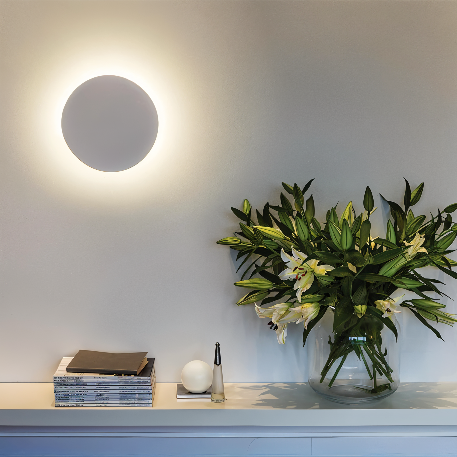 Modern wall light fixture on a neutral wall with a vase of flowers and books on a surface.