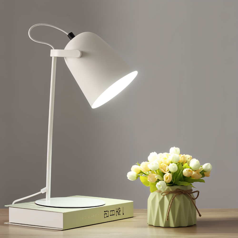 White desk lamp on a book next to a vase of flowers on a wooden surface with a gray background