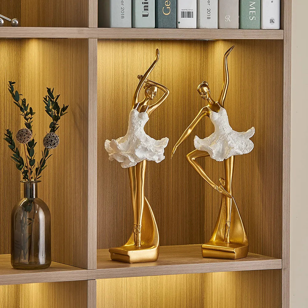 Two gold and white dancer statues on a wooden shelf with books and a vase.