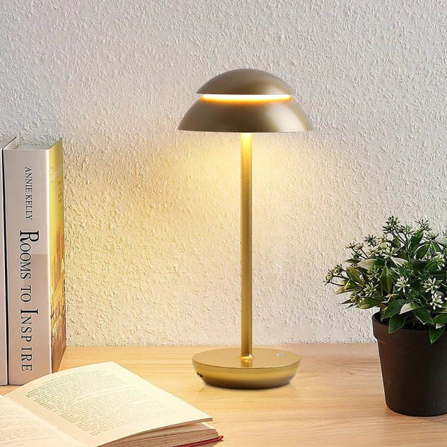 Gold table lamp on a wooden surface with a book and plant in the background
