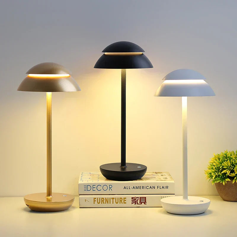 Three table lamps in gold, black, and white on a surface with books and a plant in the background.