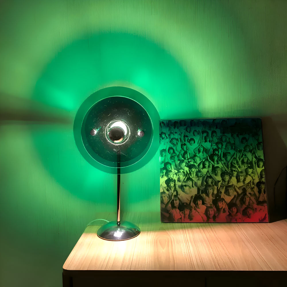Small lamp on a wooden surface with a green light casting a shadow on a wall.
