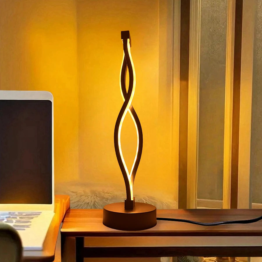 Modern table lamp with a wavy design on a desk next to a laptop.