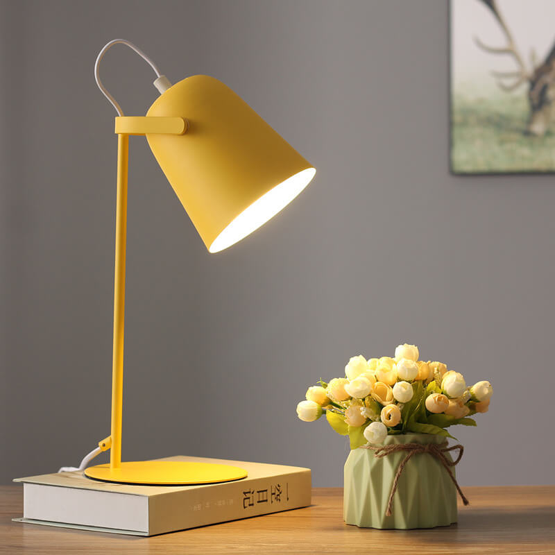 Yellow desk lamp on a wooden surface with a vase of flowers and a book against a gray wall.