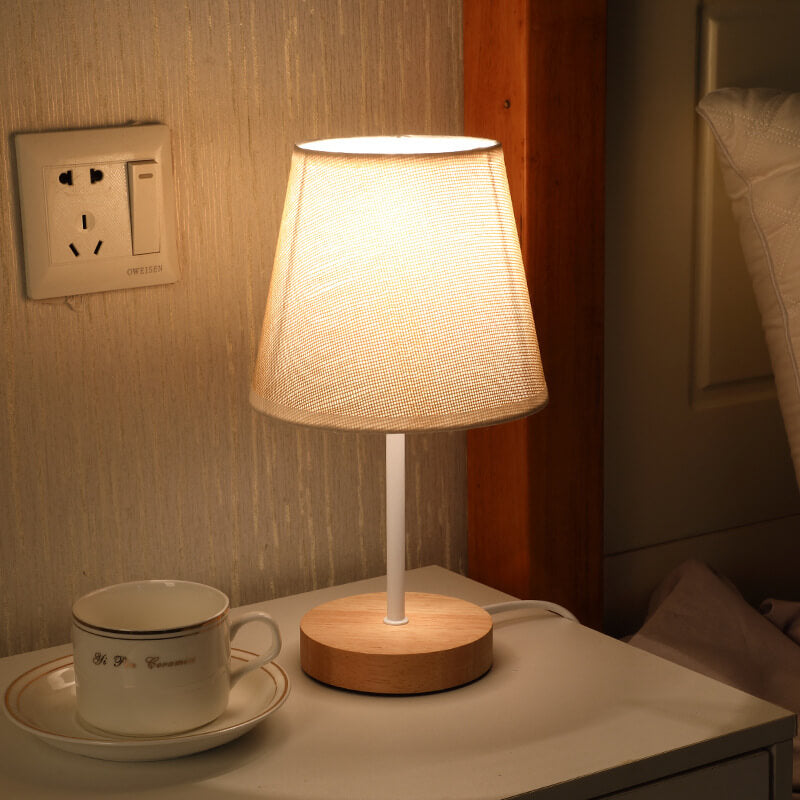 Table lamp with wooden base and white shade on a nightstand next to a cup and saucer.