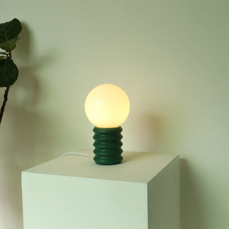 Table lamp with a green base and glowing bulb on a white pedestal against a light background
