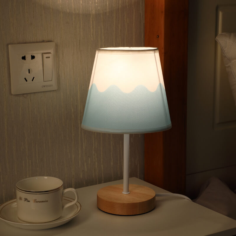 Table lamp with wooden base and mountain design on shade, next to a cup and saucer on a nightstand.
