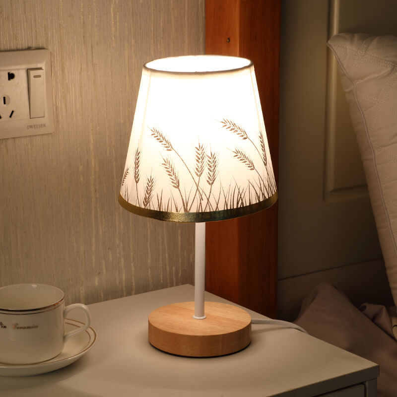 Decorative table lamp with wooden base and white shade featuring plant design, placed on a surface next to a cup.