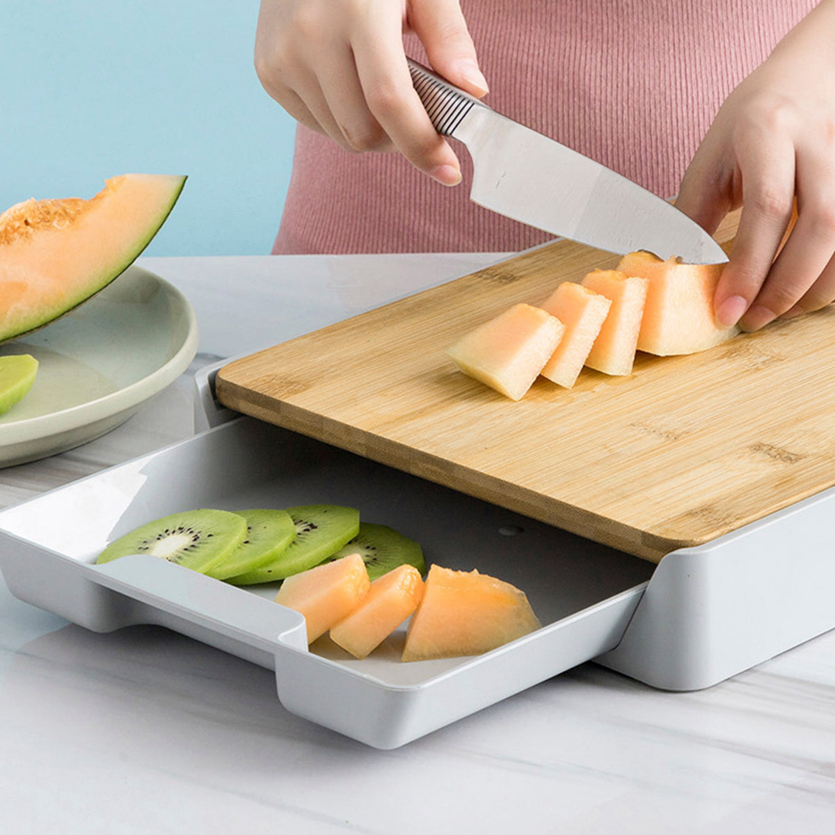 Person slicing melon on a wooden cutting board with a white tray below containing sliced fruits.