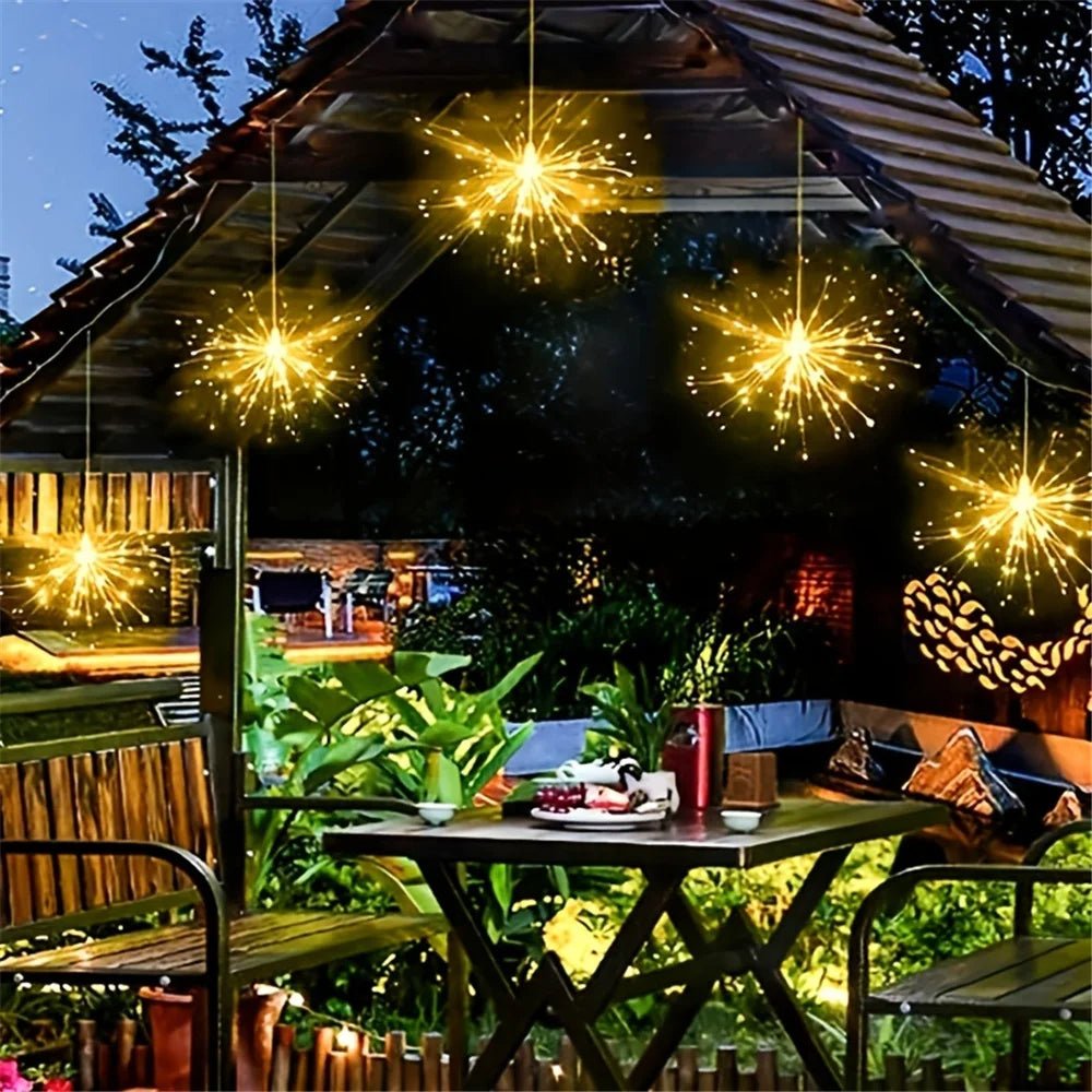 Outdoor setting with hanging string lights over a table and chairs, surrounded by greenery.