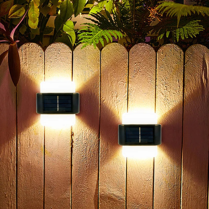 Two solar-powered outdoor wall lights on a wooden fence with plants in the background.