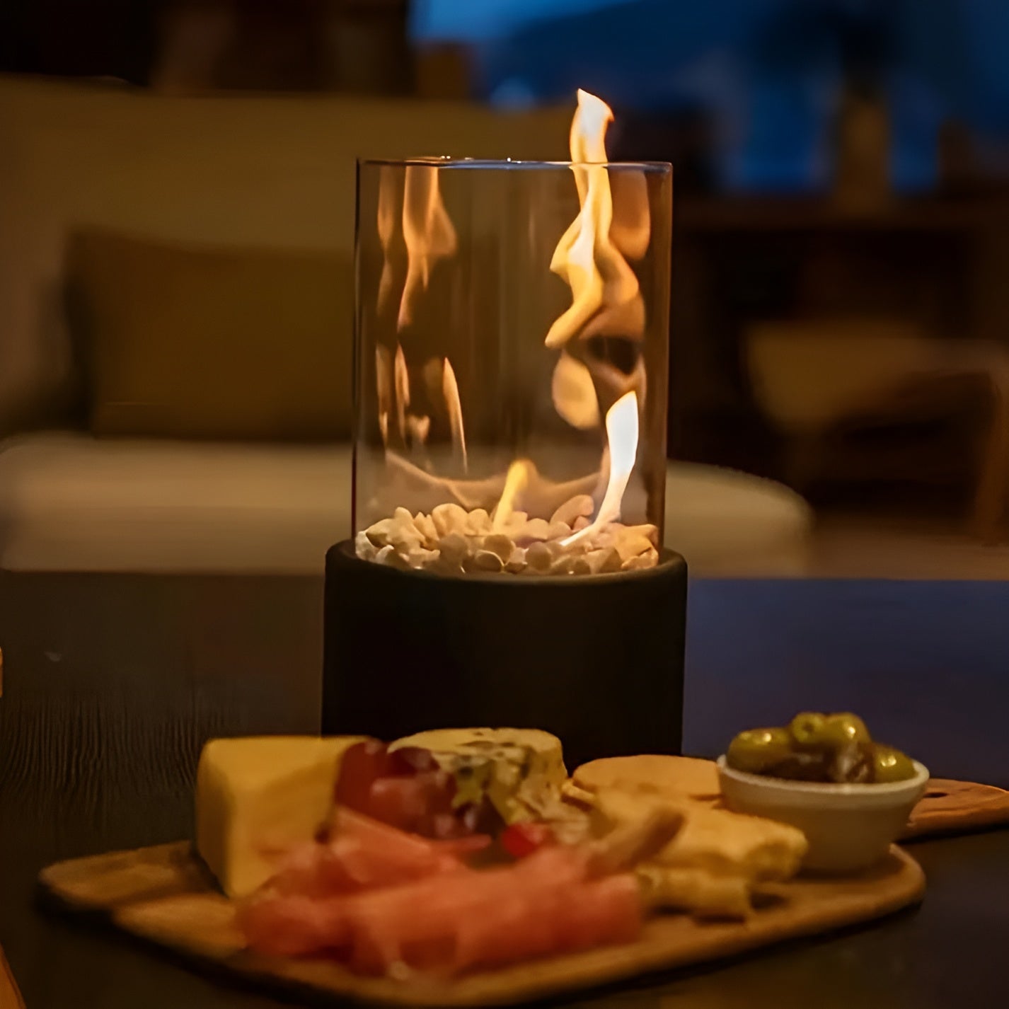 Table with a small indoor fire pit, cheeseboard, and snacks in a cozy setting.