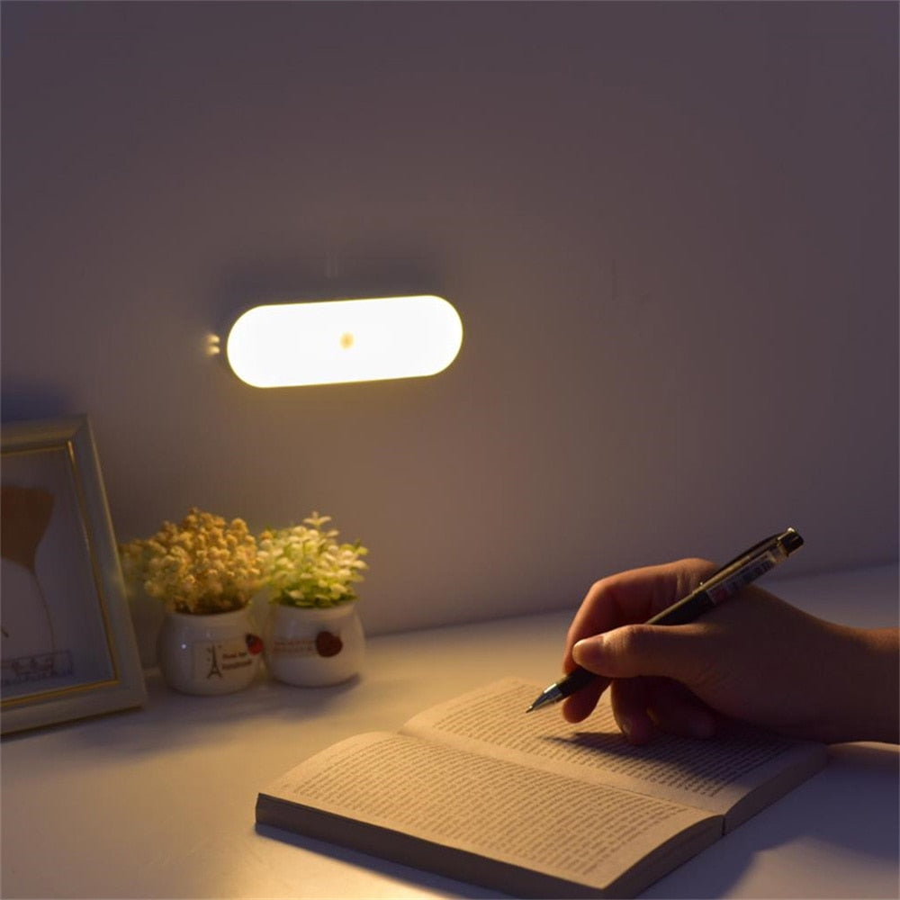 Person writing in a notebook with a pen on a table next to a small plant and framed picture, illuminated by a wall-mounted light.