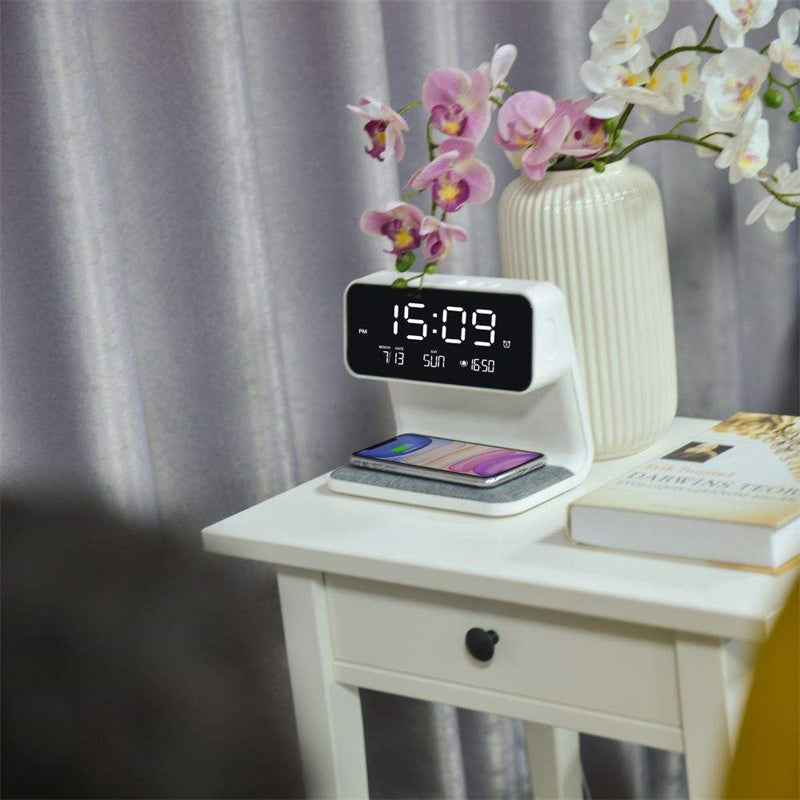 Digital clock with wireless charging pad on a nightstand with flowers and books.