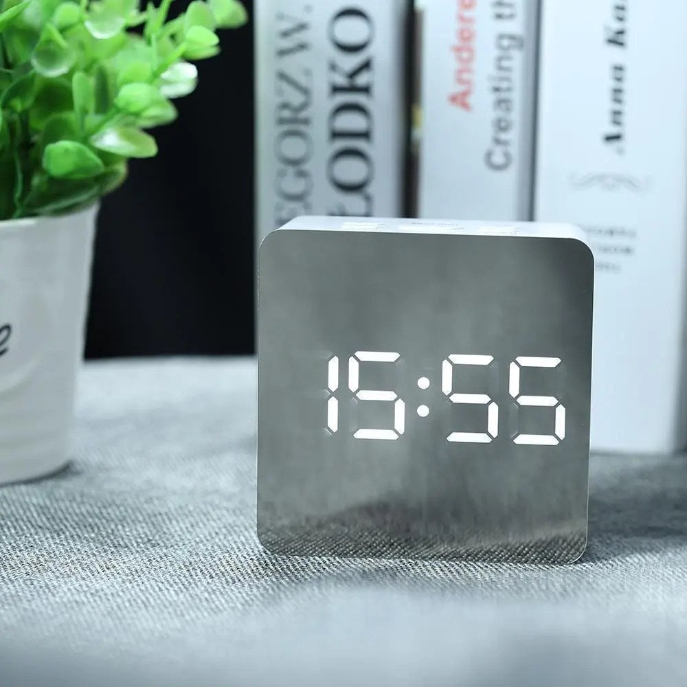 Digital clock displaying time on a table with books and a plant in the background