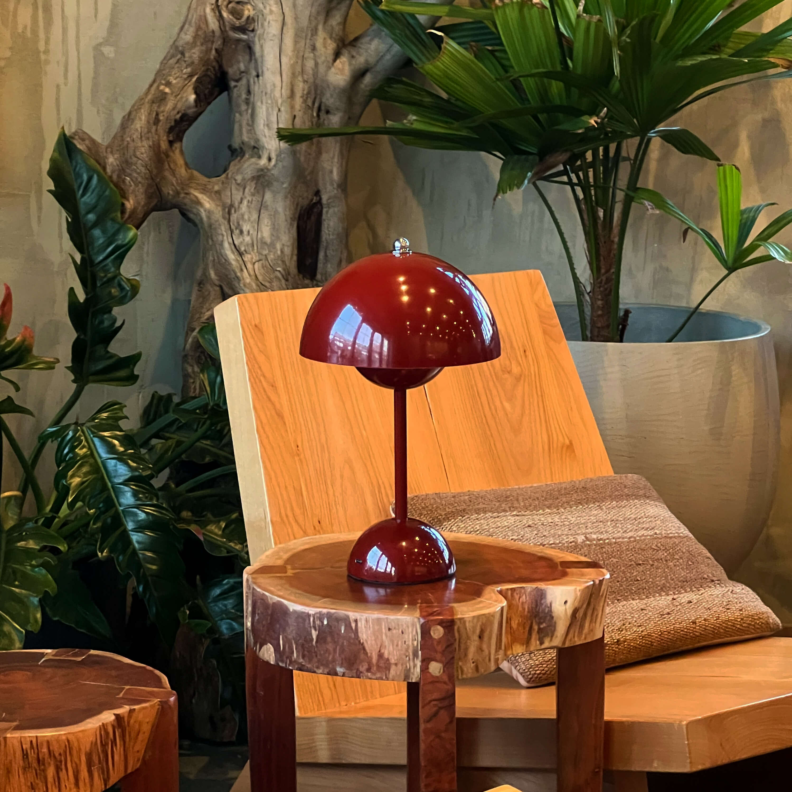 Red table lamp on a wooden stool with plants and a chair in the background