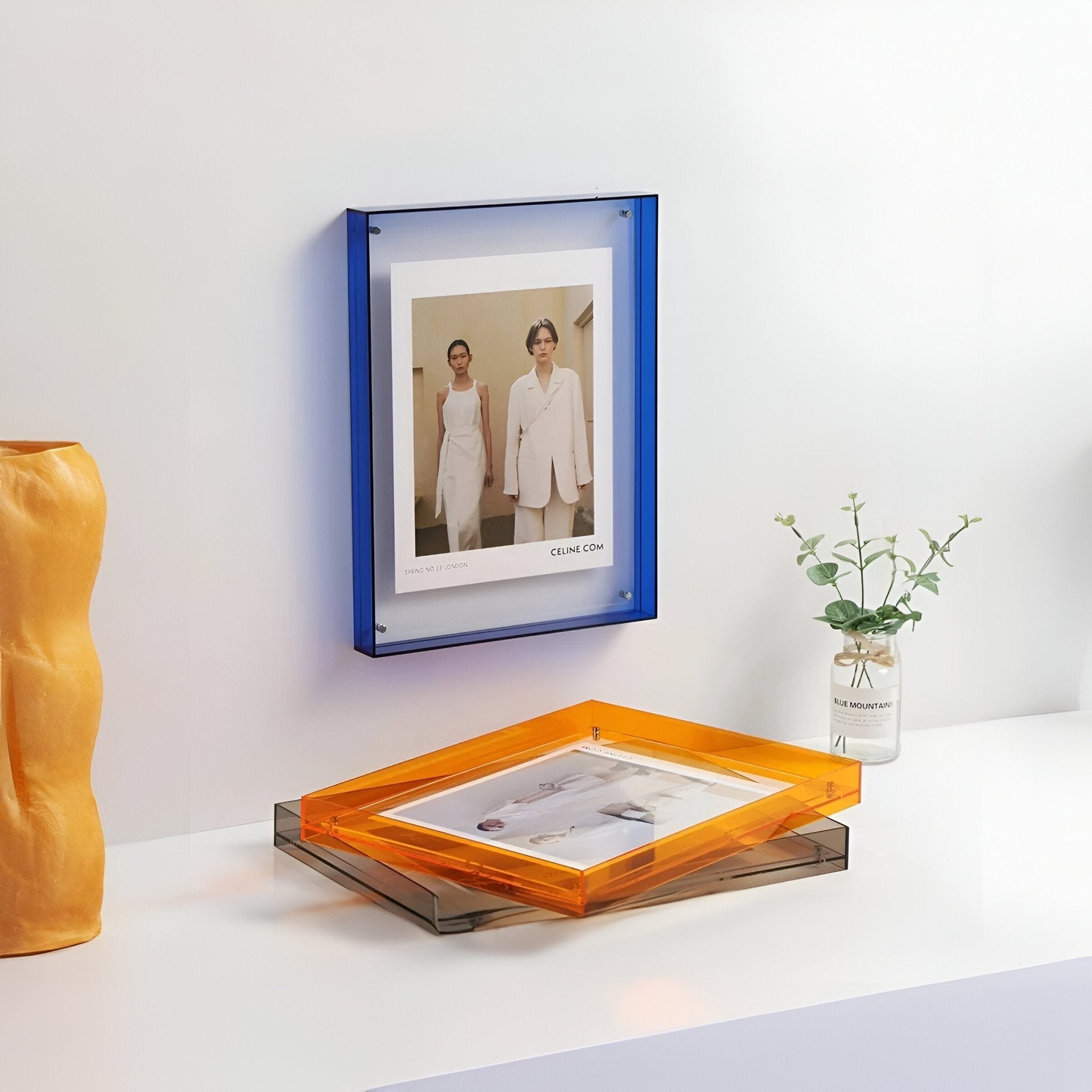 Framed photo on a wall with an orange and clear acrylic tray below, yellow vase to the left, and small plant in the corner.