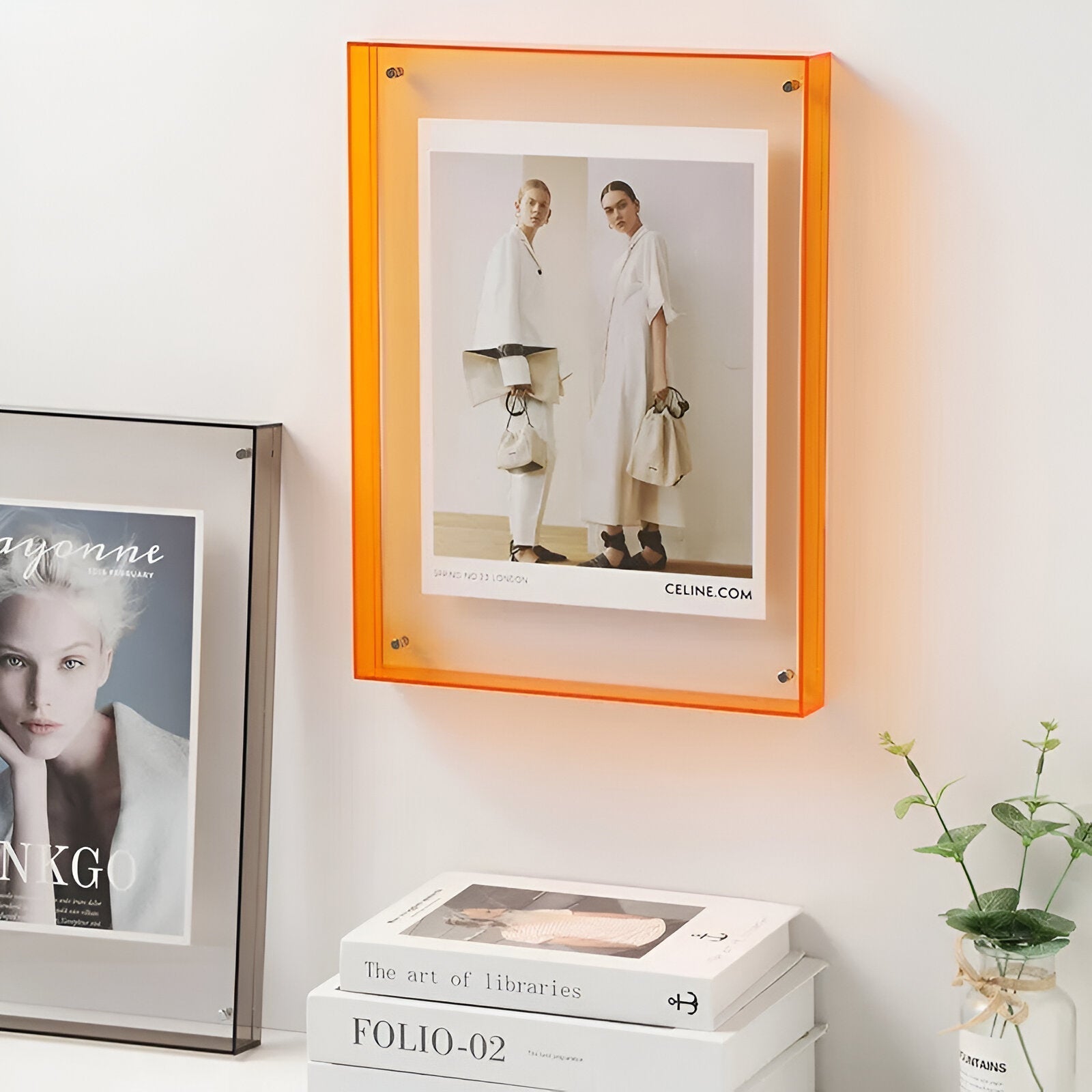 Framed picture of a fashion model with an orange frame on a wall with books and a plant.