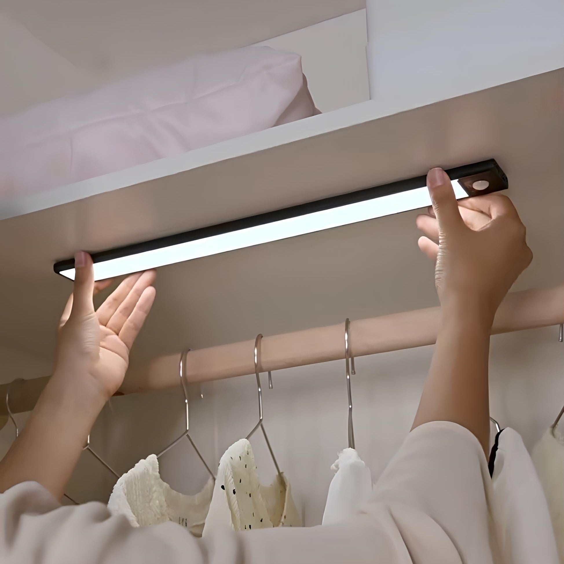 Person installing a LED strip light under a bed with clothes hanging below.