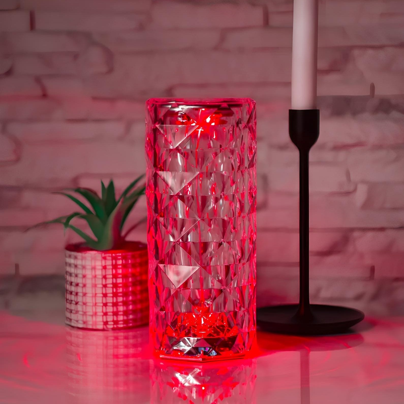 Red glass vase with geometric pattern on a reflective surface, with a plant and candle holder in the background.