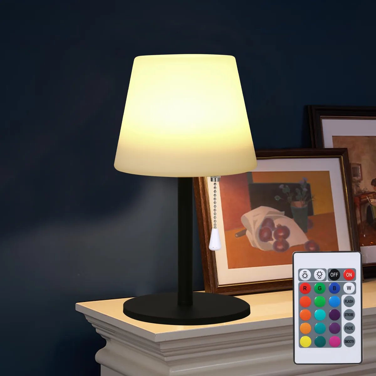 Table lamp with remote control on a surface against a dark wall with framed pictures.