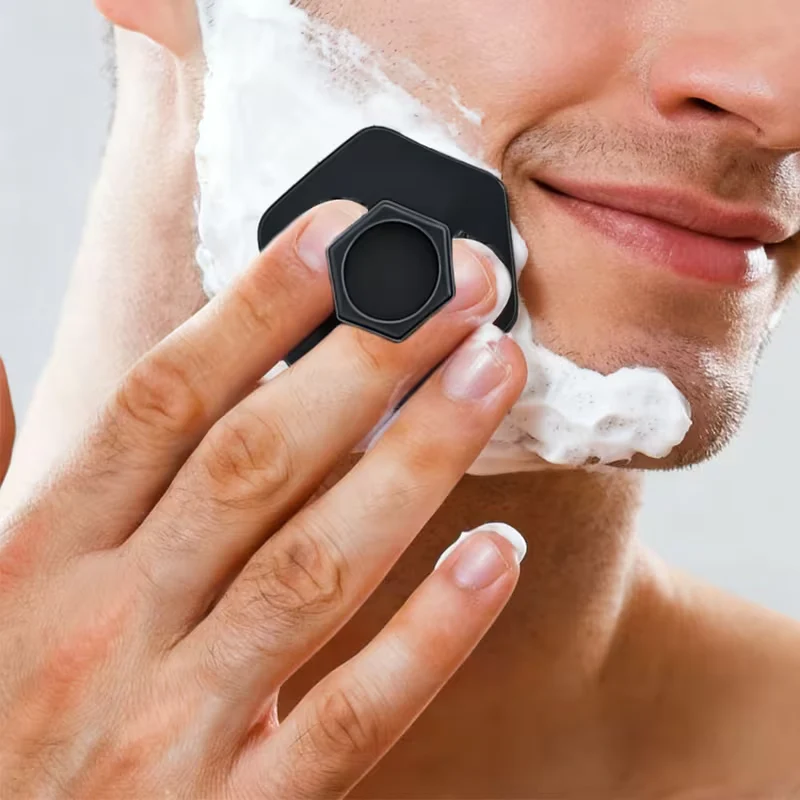 Person using a black hexagonal shaving brush with foam on a gray background