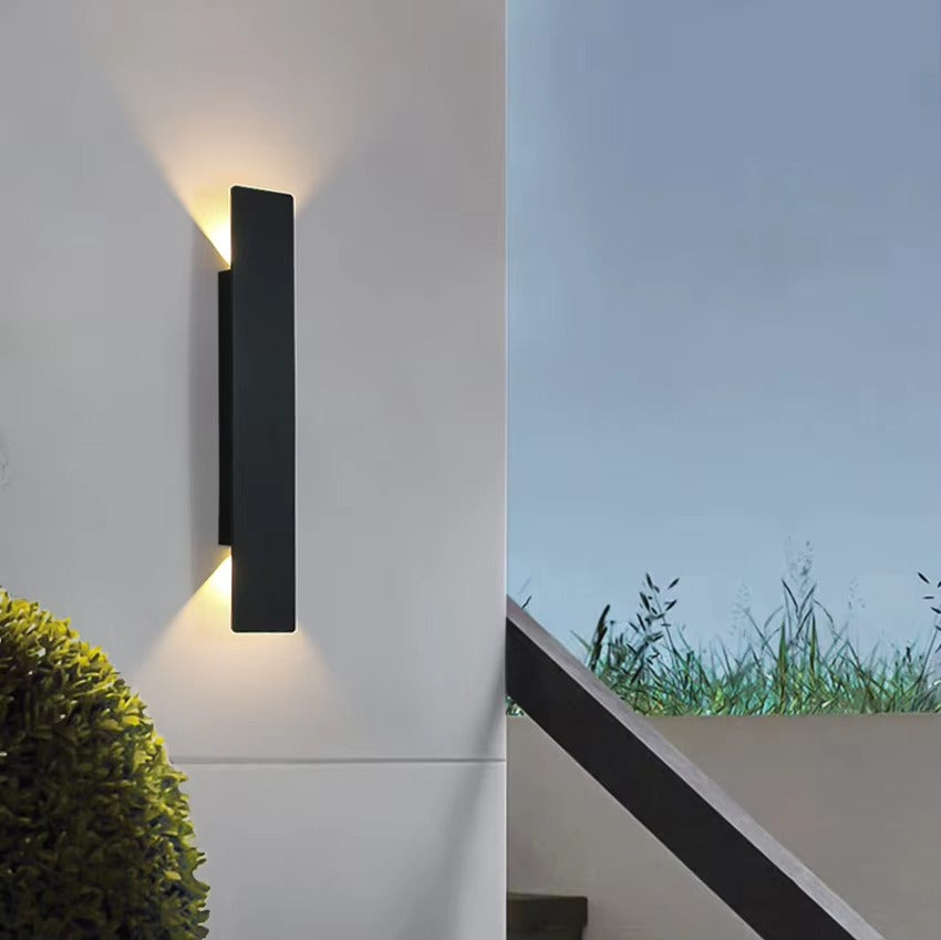 Modern outdoor wall light fixture on a white wall with a view of grass and sky.