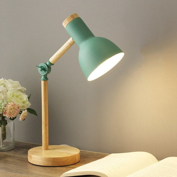 Green and wooden desk lamp on a surface with a book and flowers in the background.